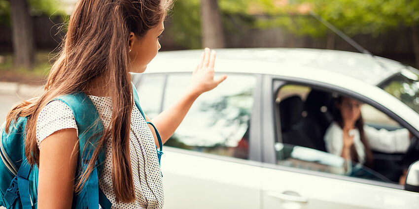 girl with backpack is dropped off at school and waves goodbye to parent in car