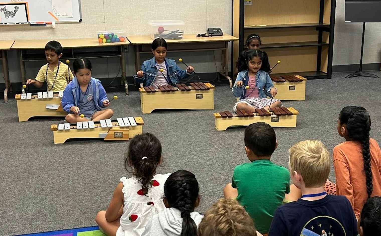 4 young students sitting on the floor playing xylophones while their classmates watch and listen